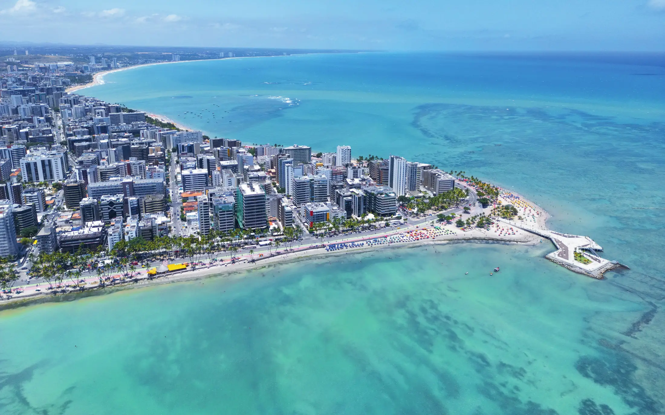 Vista aérea de una ciudad costera, con edificios junto a la playa, arrecifes y un mar turquesa.